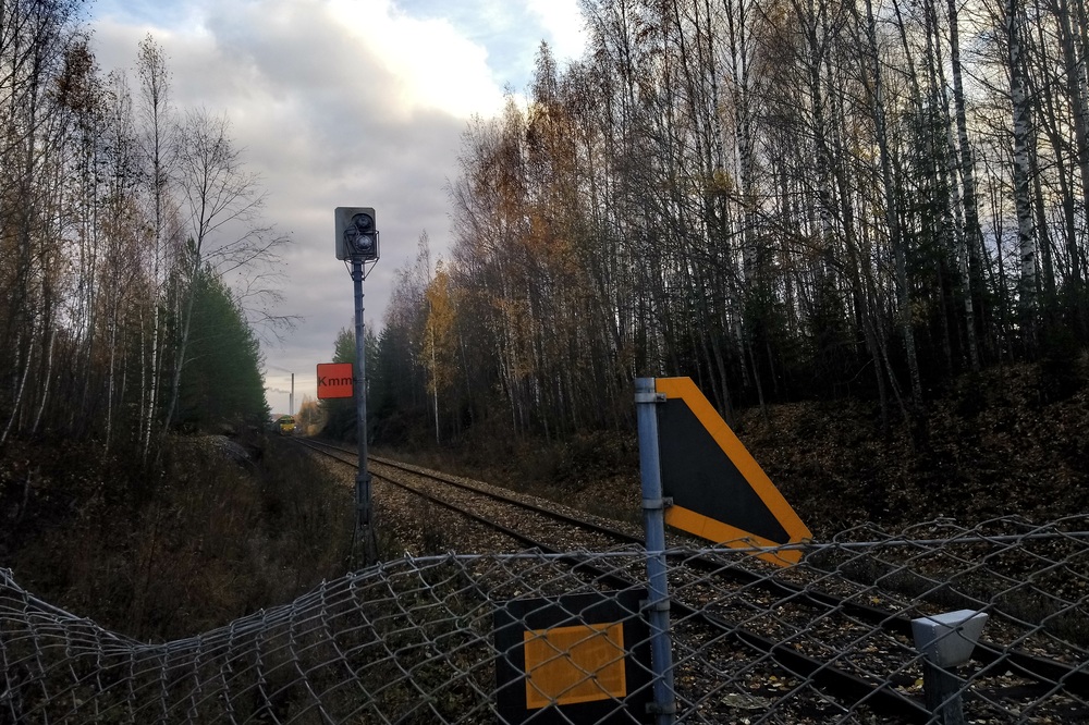 Kommilan sisäänkäynti 24.10.2019
Näkymä aidan takaa Kommilaan, Päivärinteen tasoristeyksen portilta katsottuna. Taustalla näkyy Stora Enson Tve4 järjestelmässä vaunuja Kommilan ratapihalla. Veturin syrjäytti pian tämän jälkeen samana päivänä toimitettu Dr35.
